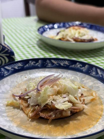 A sopito (beef, lettuce, cheese, red onion and broth over a tostada) on a blue-rimmed plate, sitting on a green and white checkerboard tablecloth