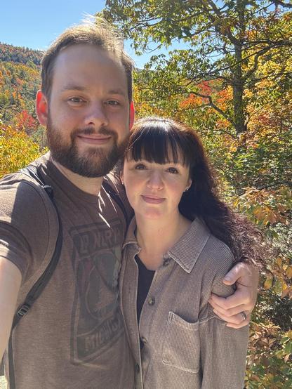A man and woman smiling together outdoors during autumn with colorful fall foliage in the background.