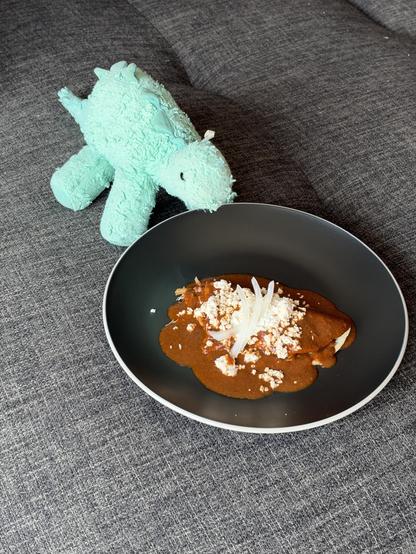 A blue-green stuffed stegosaurus sits in front of a plate with an enchilada covered in a brown sauce with cheese and onion on top