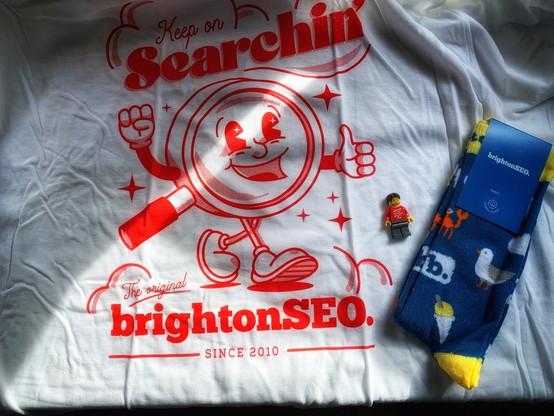 White BrightonSEO tee shirt with keep on searchin’ and and mascot in red with the original Brighton SEO since 2010 on the base. Resting on the tee shirt is also a Lego minifig with a red tee shirt with the same as the real tee shirt in white and also to the side a pair of blue brightonSEO socks with seagulls and ice creams on. 