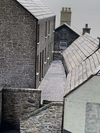 View down an alleyway between a row of stone cottages and the roofs of building in front of them at a lower level. At the end is the upstairs side window of another house just over the end wall. In the foreground is a set of steps coming down between retaining walls. It is a model.