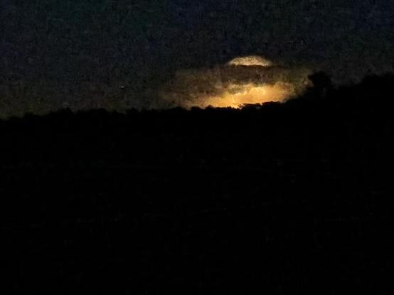 Dark night photo with horizon across middle of image with bright orange lightning showing up a cloud 140 miles away.
