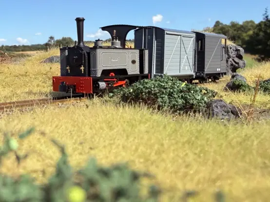 Small light gray narrow gauge locomotive pulling a medium gray closed van and a darker gray guards van  out from a rocky cutting through some straw coloured grassland.