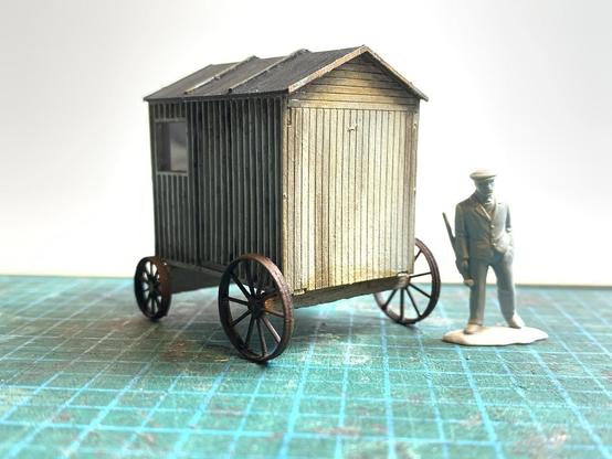 Model of an old shepherds hut with a plastic figure alongside for scale, on a cutting mat.